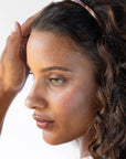 France Luxe thin headband in a warm caramel and amber color, pictured holding curly, brown hair in a loose hairstyle.