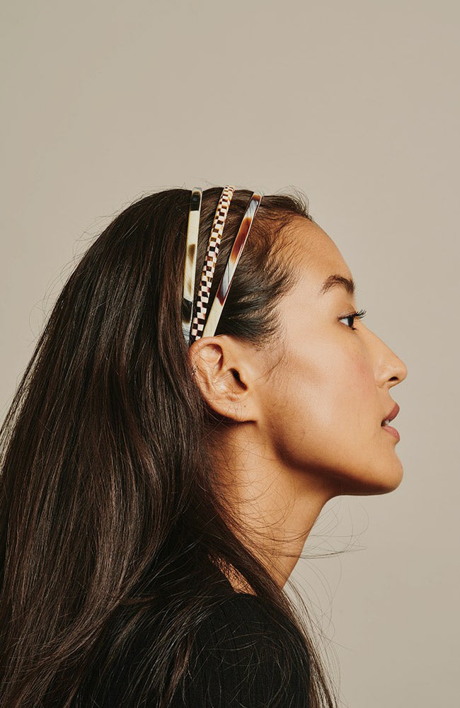 France Luxe thin headband in a Natural Horn, Black Horn, and Damier color, pictured holding straight, black hair in a loose hairstyle.