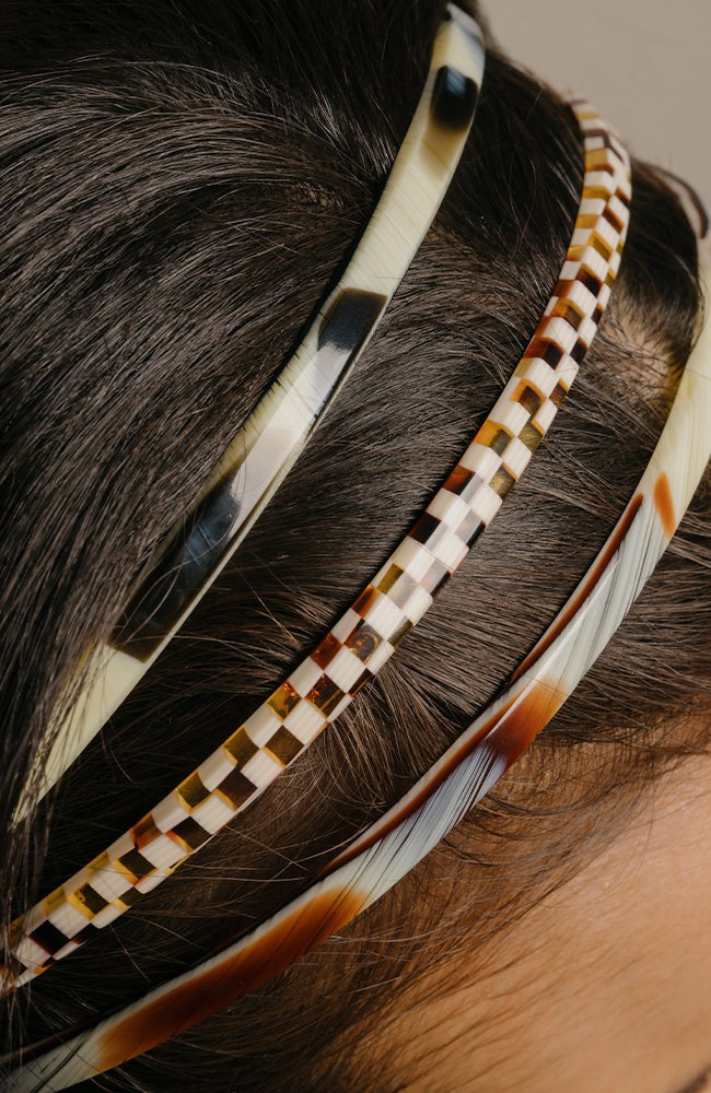 France Luxe thin headband in a Natural Horn, Black Horn, and Damier color, pictured holding straight, black hair in a loose hairstyle.