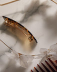 Classic Tortoise French Barrette, with a white flower headband and wide tooth comb on a counter.
