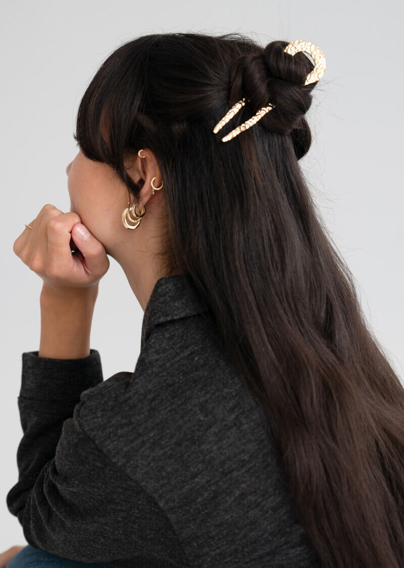 Person with styled, dark brown hair using a hammered, gold hair pin to hold a half up bun.