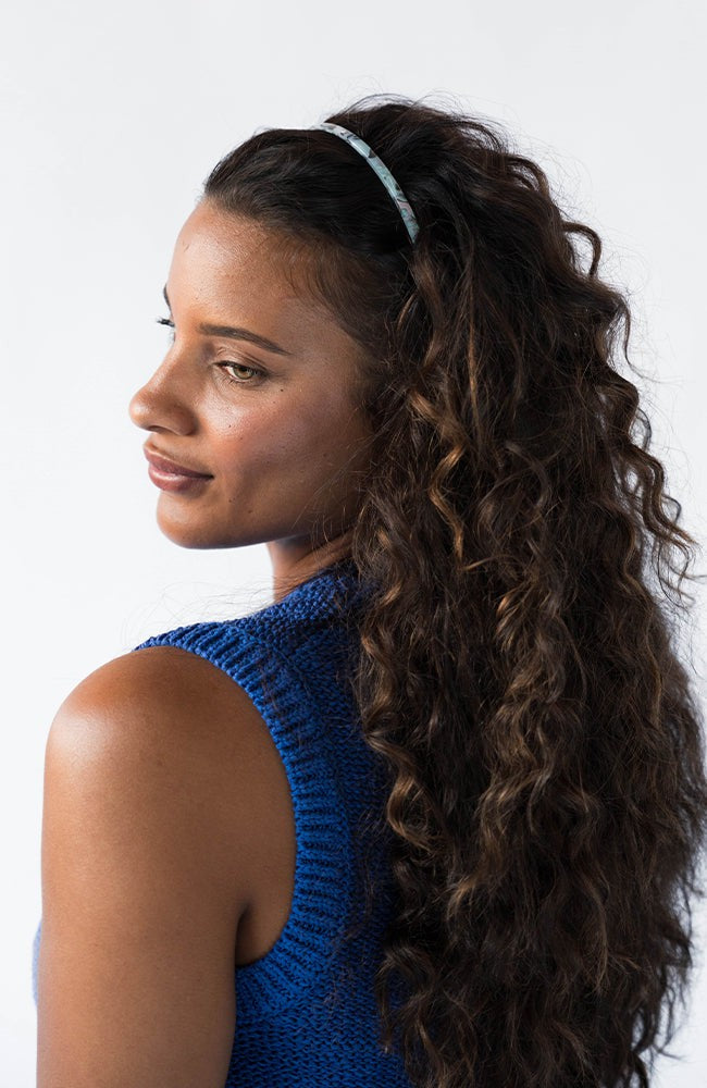 France Luxe thin headband in a light blue, faux shell color, pictured holding curly, brown hair in a loose hairstyle.
