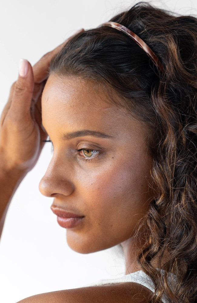 France Luxe thin headband in a warm caramel and amber color, pictured holding curly, brown hair in a loose hairstyle.