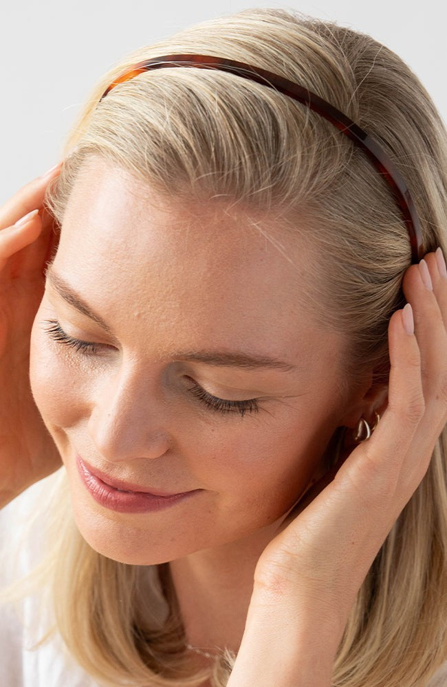 France Luxe thin headband in a rich amber and mahogany brown color, pictured holding straight, blonde hair in a loose hairstyle.