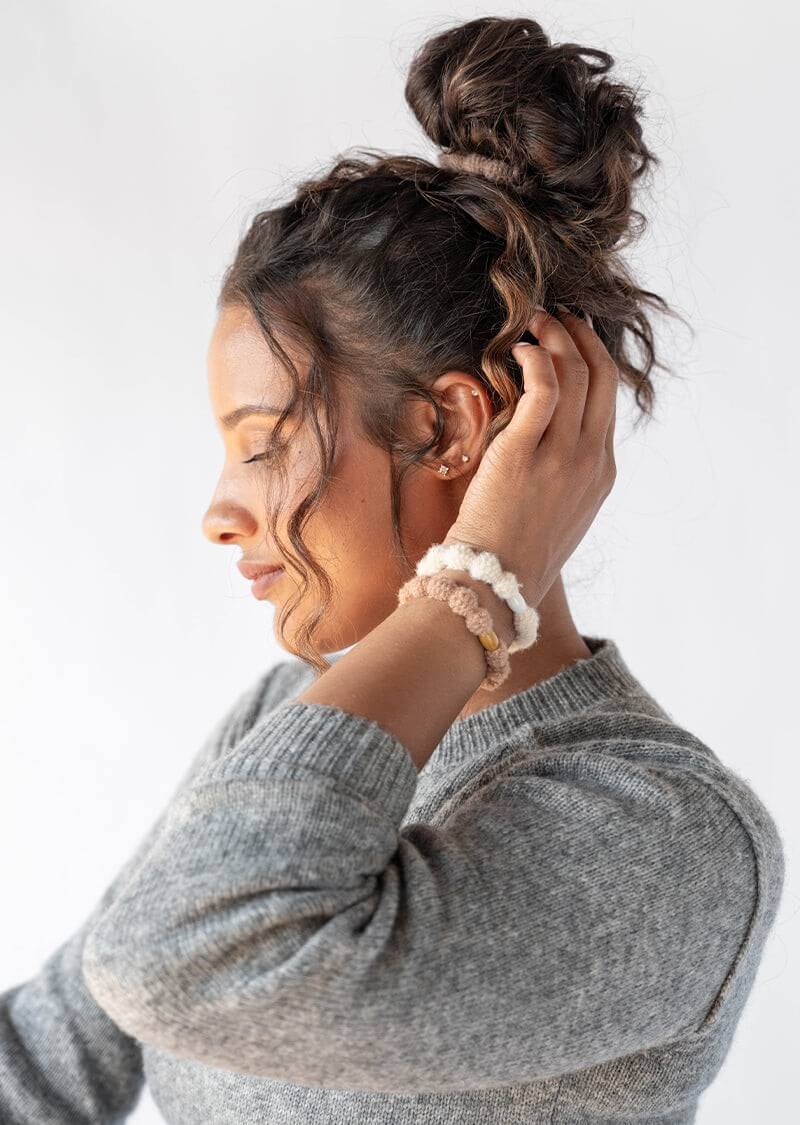 A woman with curly brown hair in a loose bun held with a cute L. Erickson hair tie, with two bubble ponies on her wrist
